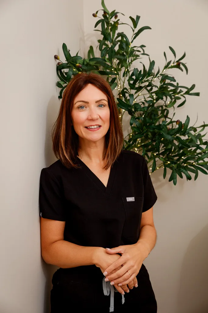 Woman with reddish-brown hair wearing black scrubs, smiling at camera with hands clasped, standing in front of a potted plant.