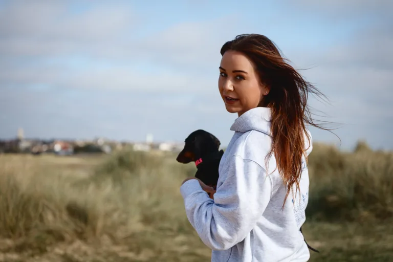A woman in a light blue jacket holds a small black dog while looking over her shoulder in an open field.