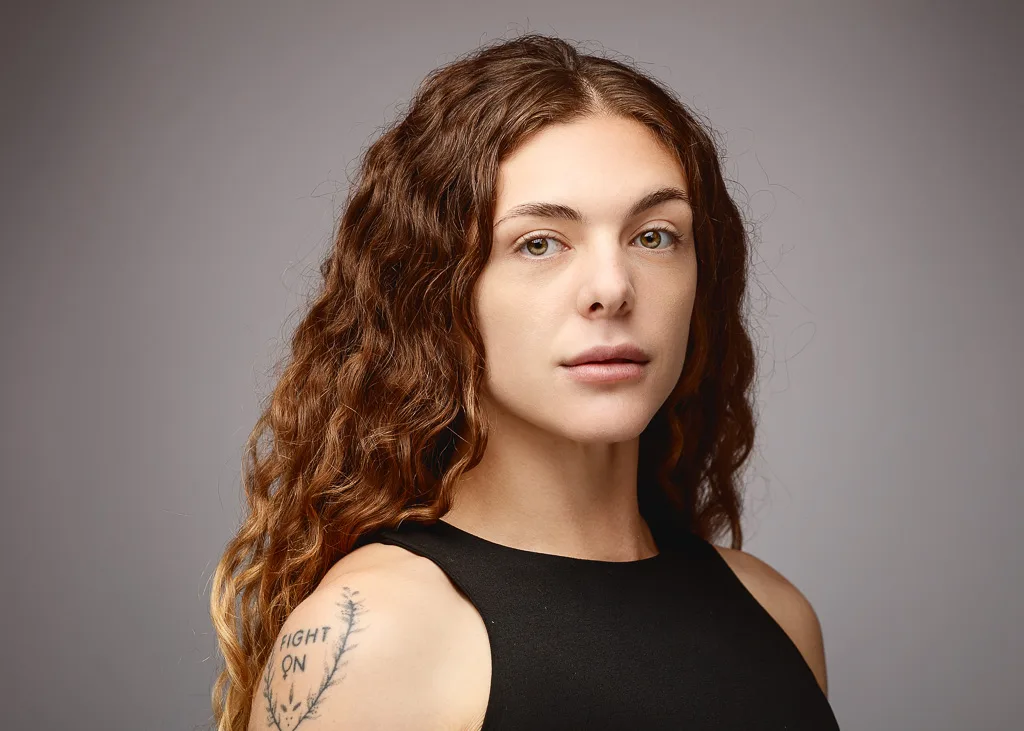 A person with long, curly reddish-brown hair wearing a black sleeveless top, photographed against a grey background. They have a tattoo visible on their shoulder.