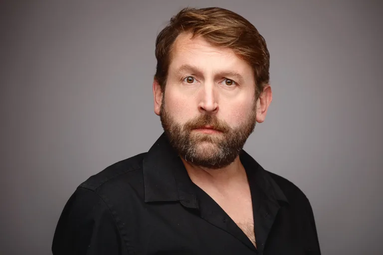 A man with brown hair and a full beard wearing a black shirt, photographed against a grey background.