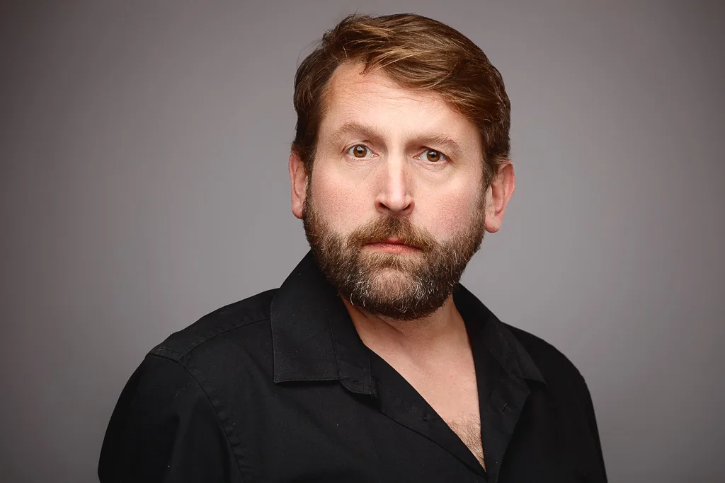A man with brown hair and a full beard wearing a black shirt, photographed against a grey background.