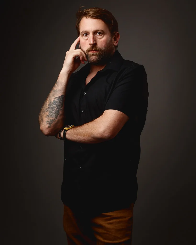 A man with a beard and brown hair wearing a black shirt and brown trousers stands against a grey background, hand to his face in a thoughtful pose.