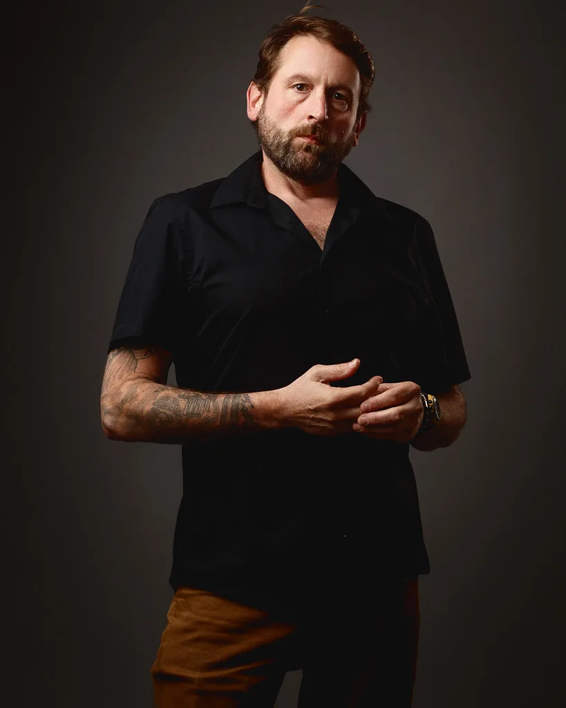 A bearded man in a black short-sleeved shirt and brown trousers stands against a grey background, gesturing with his hands while looking to the side.
