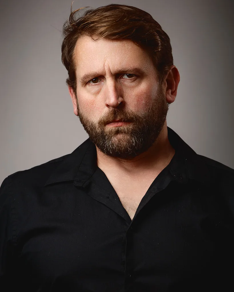 A man with a full beard and brown hair wearing a black shirt, photographed against a neutral grey background.