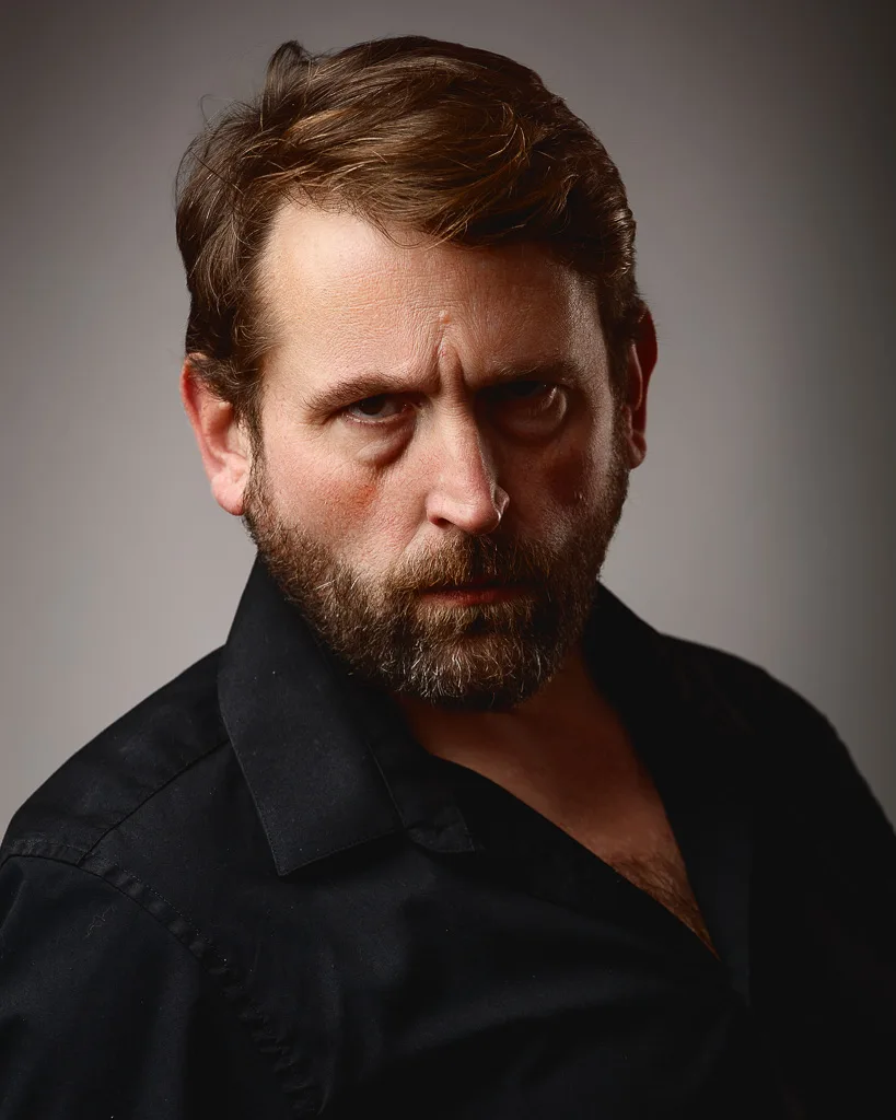 Portrait of a bearded man with brown hair wearing a black shirt against a neutral grey background, looking downward.