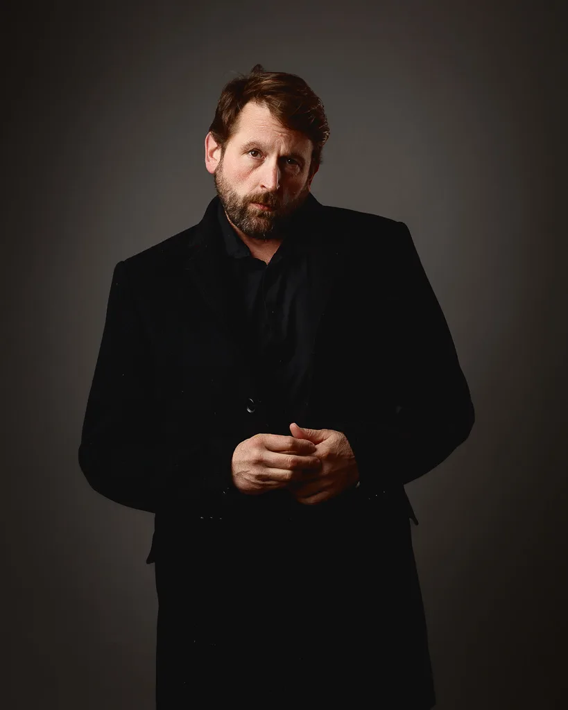 A man with brown hair and a beard wears a black blazer and shirt against a neutral grey background, looking slightly to the side.