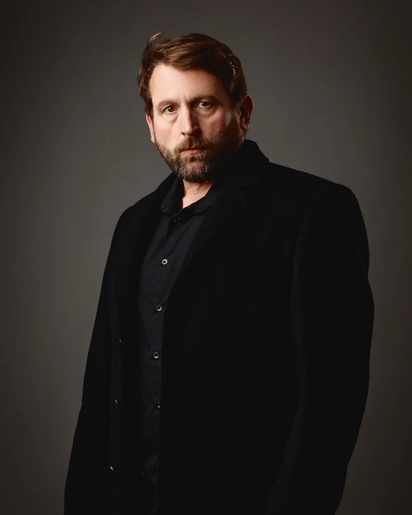 A man with brown hair and a beard wears a black jacket and shirt against a neutral grey background, looking to the side.