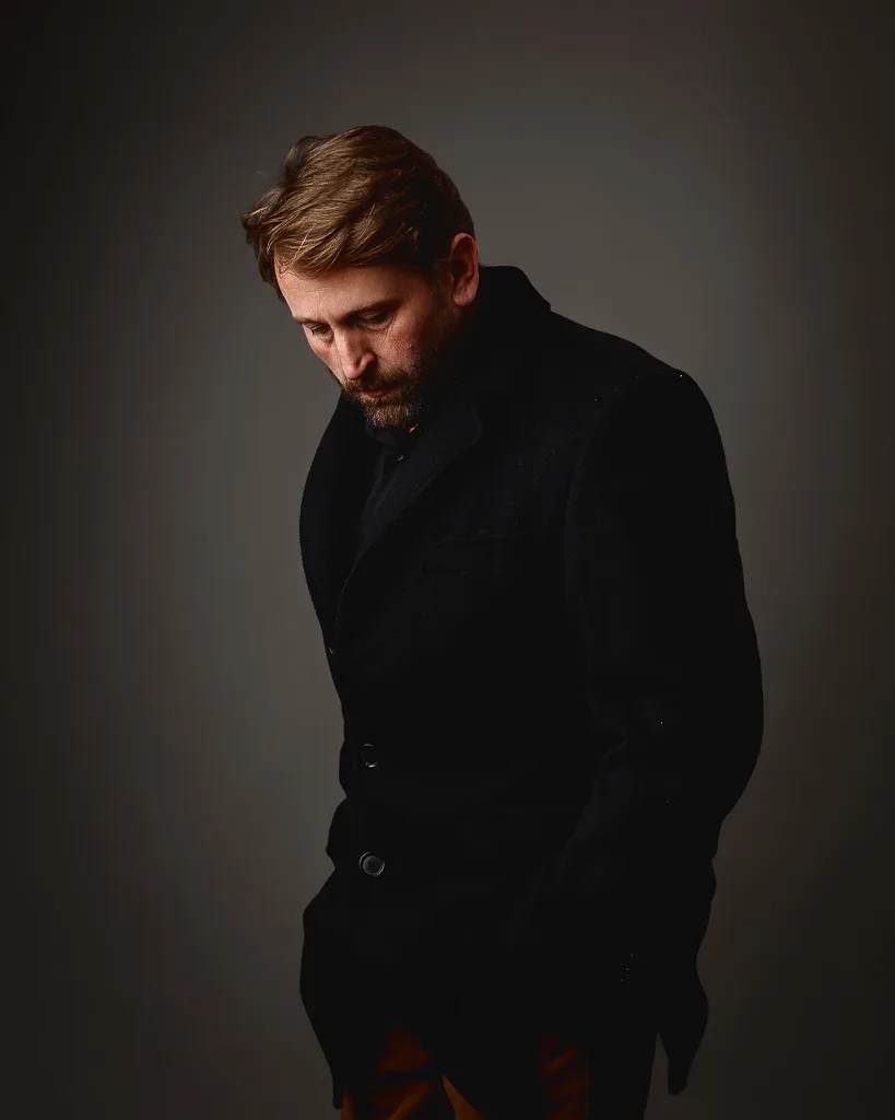 A man with a beard wearing a black blazer stands against a grey background, looking downward with a contemplative expression.