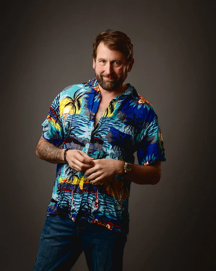A bearded man wearing a vibrant tropical print shirt and jeans stands against a grey background, smiling at the camera.