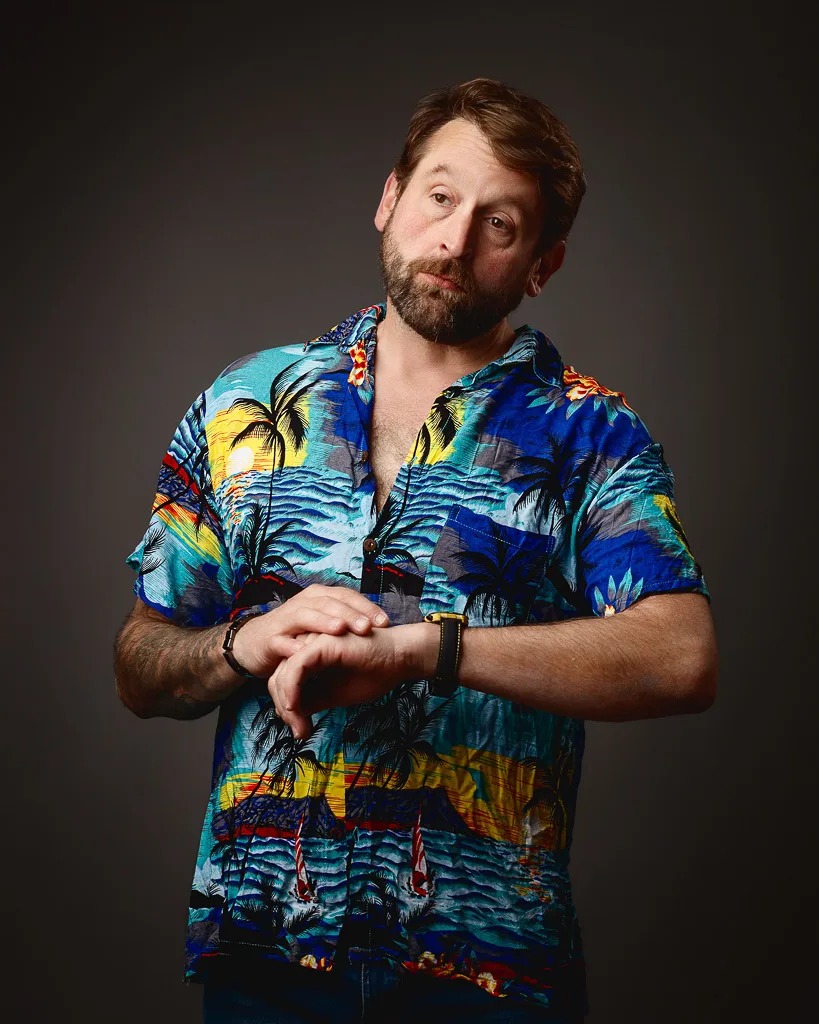 A man with brown hair and beard wearing a colourful tropical print shirt looks at his watch against a dark grey background.