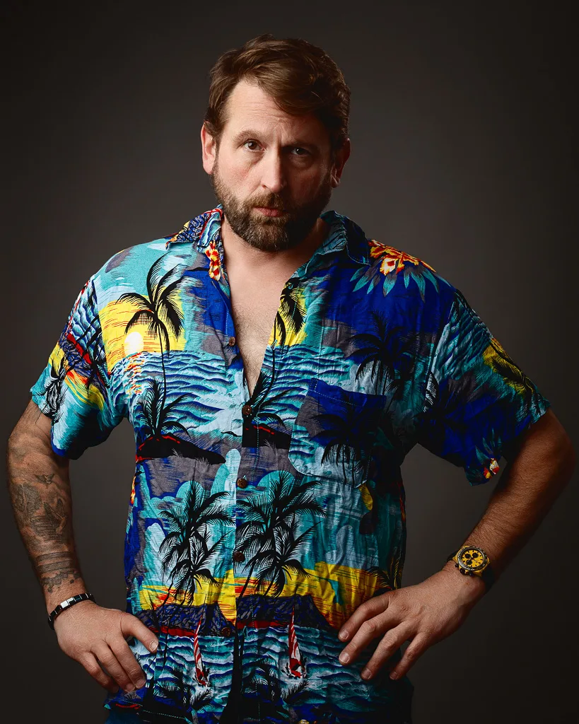 Man with beard wearing a vibrant tropical print shirt with palm trees and ocean scenes, posing against a dark grey background.
