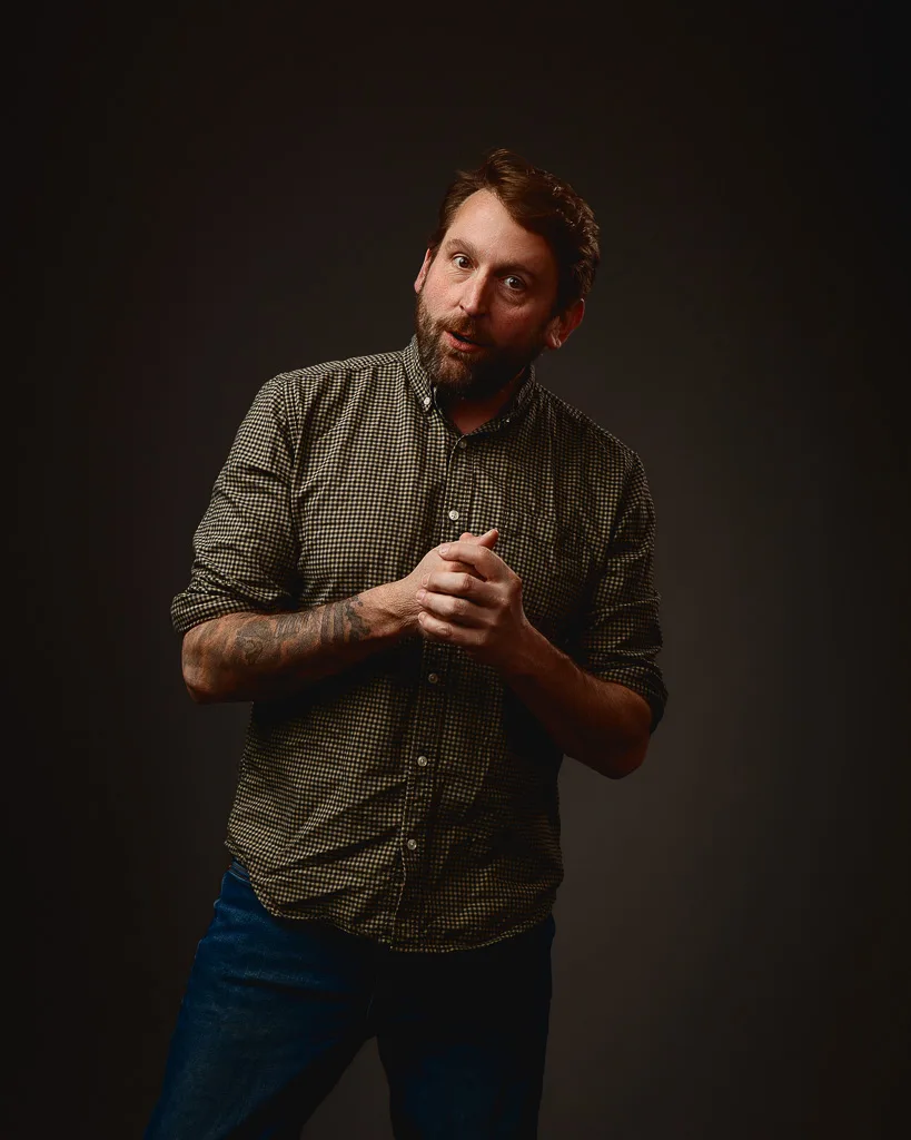 A bearded man in a checked shirt and jeans gestures while speaking against a dark background.