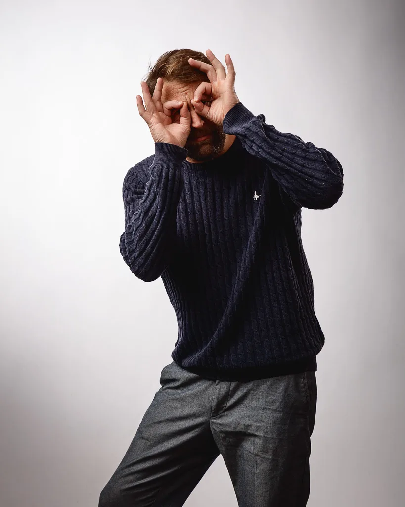 A man in a dark ribbed sweater and grey trousers shields his eyes with both hands against a plain white background.