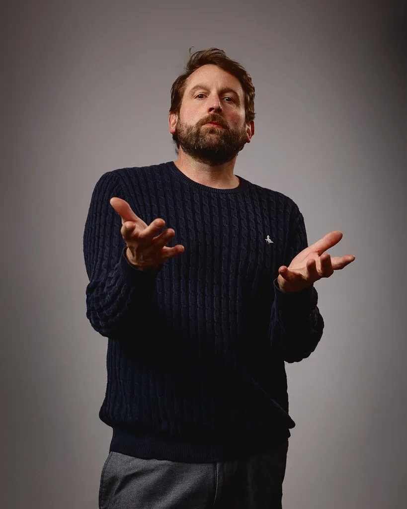 A bearded man in a dark ribbed jumper gestures expressively with both hands raised, looking upward against a plain grey background.