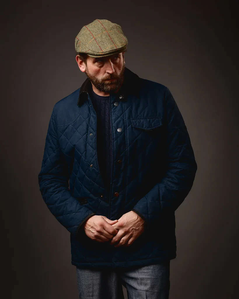 A bearded man wearing a navy quilted jacket, dark jumper, flat cap, and grey trousers stands against a dark background.