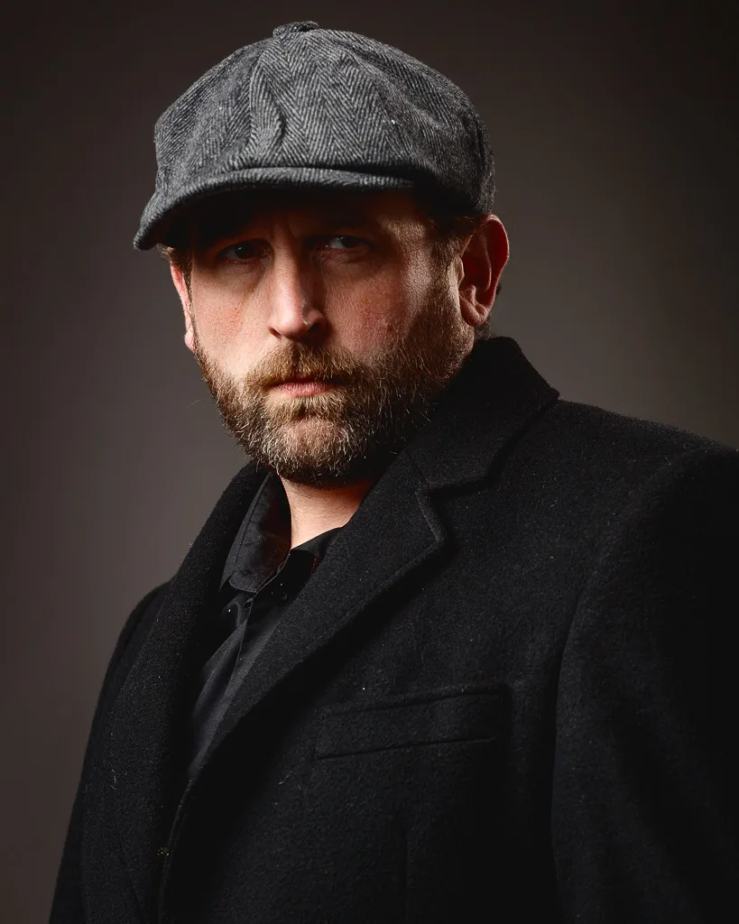 A man with a full beard wearing a grey herringbone flat cap and black coat, looking directly at the camera against a dark background.