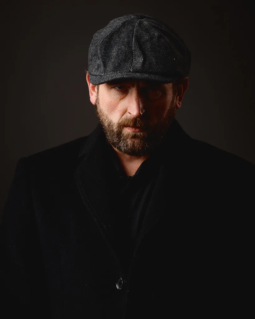 A bearded man wearing a dark coat and grey flat cap looks downward against a black background.