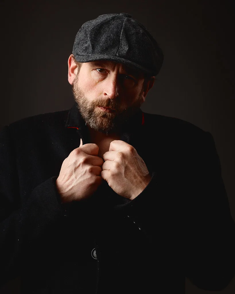 A bearded man wearing a dark flat cap and black jacket, with hands clasped at his chest, against a dark background.