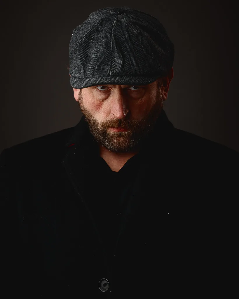 A bearded man wearing a dark flat cap and black shirt gazes downward against a dark background.