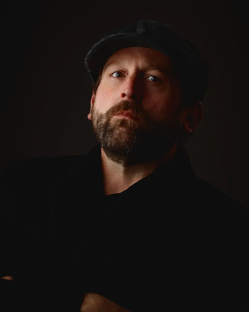 A bearded man wearing a black beanie and dark clothing is photographed against a black background, looking upward.
