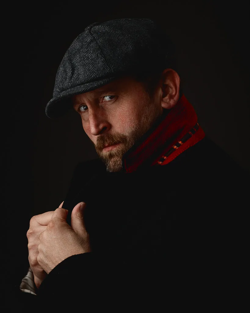 A bearded man wearing a flat cap and red scarf gestures toward the camera against a black background.
