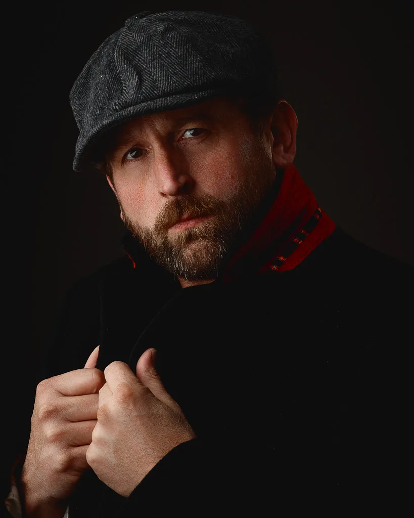 A bearded man wearing a grey herringbone flat cap and red-lined black jacket against a dark background, looking toward the camera.